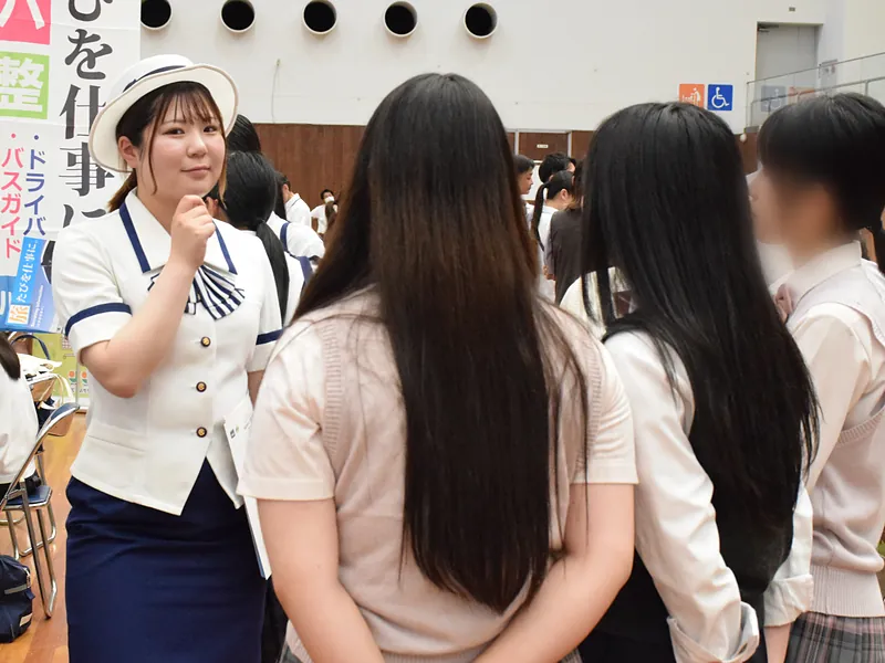 バス会社のブースで、バスガイドの制服を着た担当者が、高校生に仕事の話をしている様子